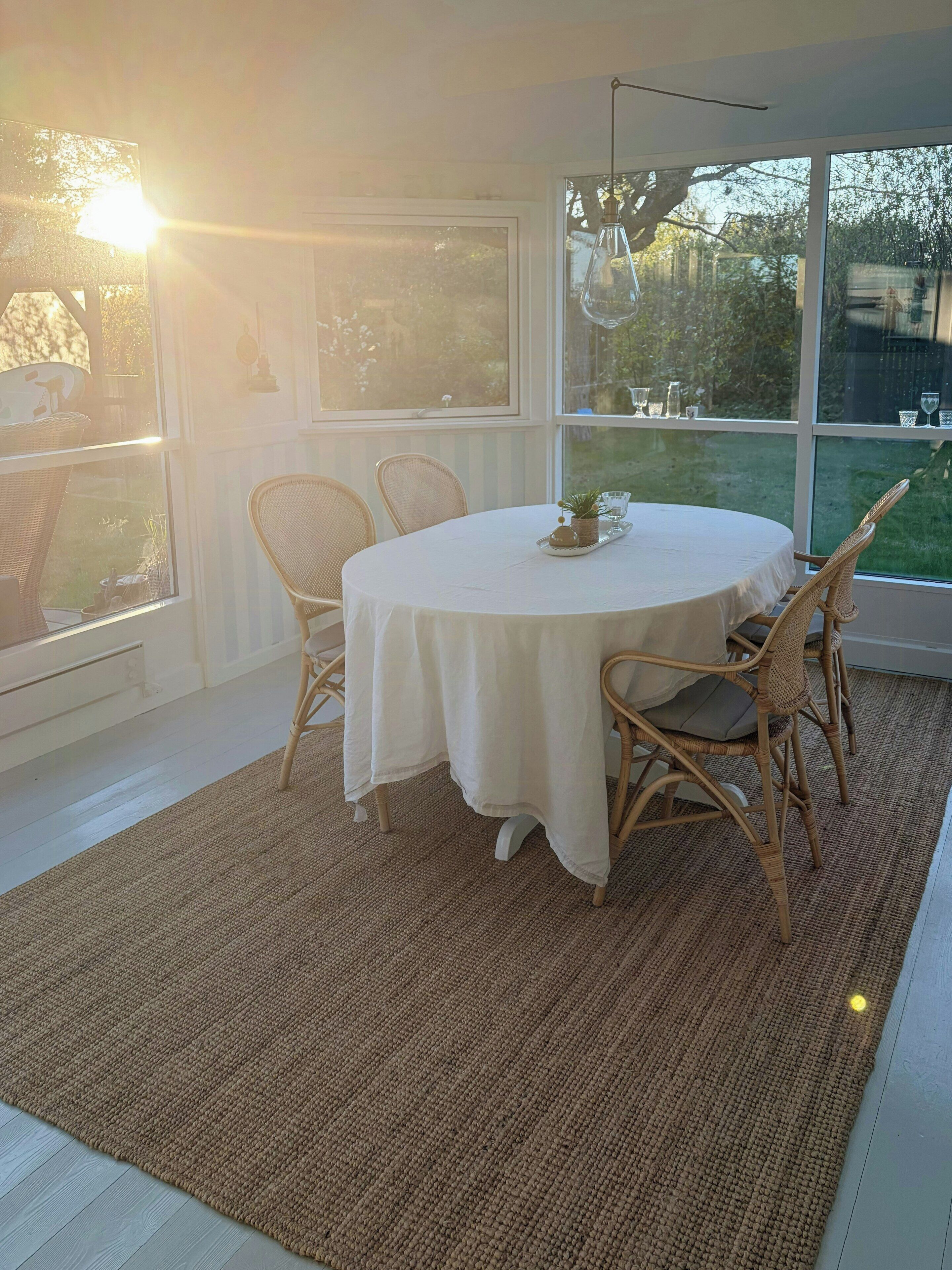 Une table ronde recouverte d’une nappe blanche, entourée de chaises en osier, dans une pièce dotée de grandes fenêtres et ornée d’un tapis tissé.
