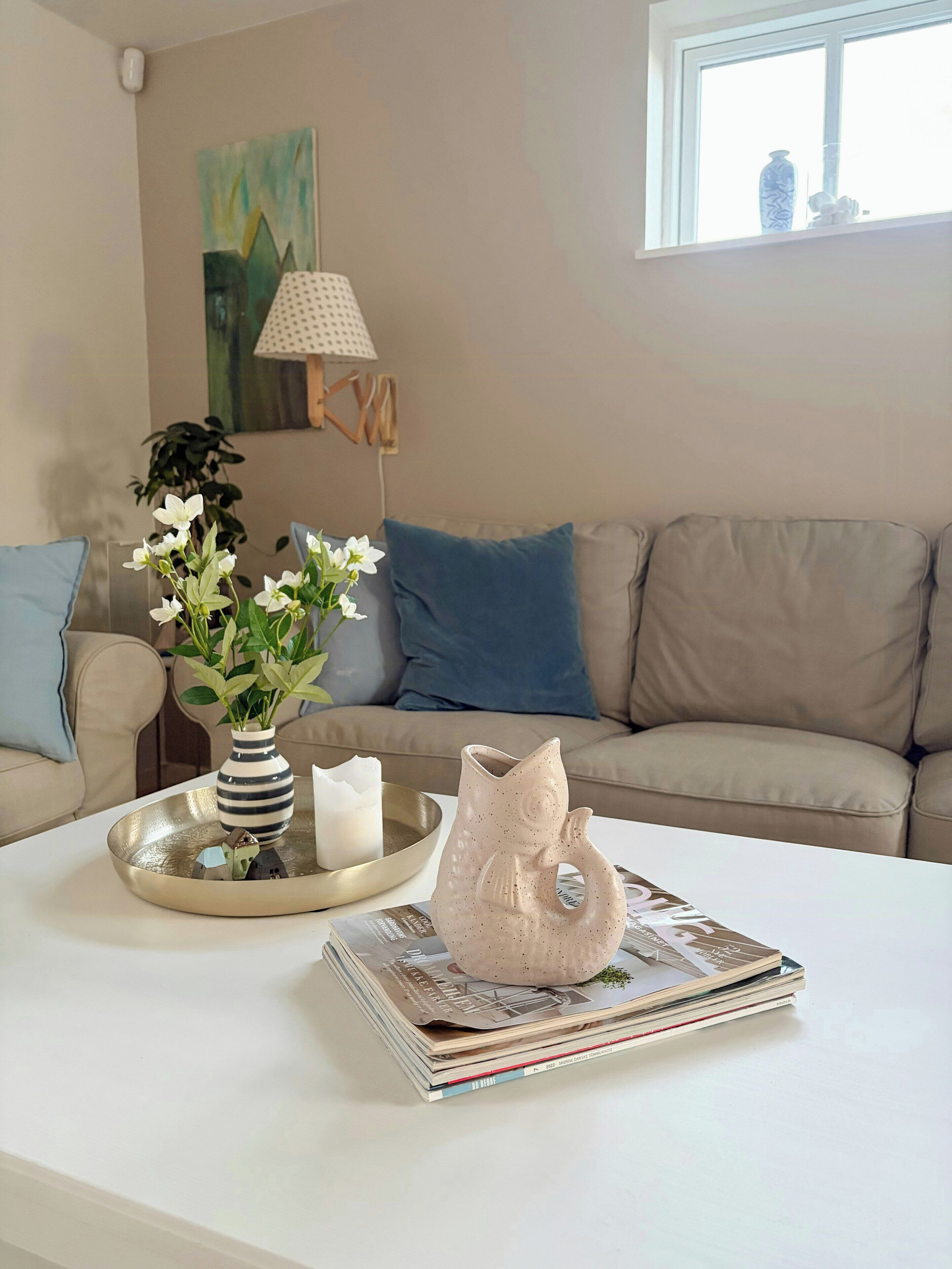 Una sala de estar con una mesa de centro blanca, un florero, un florero con forma de pez, una lámpara y un sofá con cojines.