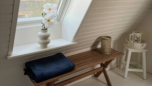 Ein Zimmer mit geneigter Decke, einem Fenster mit Blick auf Grünflächen, einer Holzbank, einem kleinen Tisch mit einem Korb und einem weißen Krug auf dem Tisch.