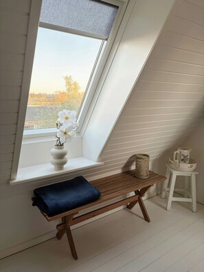 Ein Zimmer mit geneigter Decke, einem Fenster mit Blick auf Grünflächen, einer Holzbank, einem kleinen Tisch mit einem Korb und einem weißen Krug auf dem Tisch.