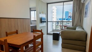 Una habitación moderna con una mesa de comedor de madera, un sofá, un televisor montado en la pared y un ventanal con vistas a edificios.