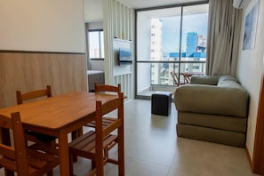 A modern room with a wooden dining table, a sofa, a TV mounted on the wall, and a large window with a view of buildings. - Cozy Studio at Enjoy Itapoã (Vila Velha)