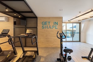 Ein Fitnessraum mit Laufbändern, Crosstrainern und einem stationären Fahrrad. An einer Wand steht der Schriftzug 'GET IN SHAPE'.