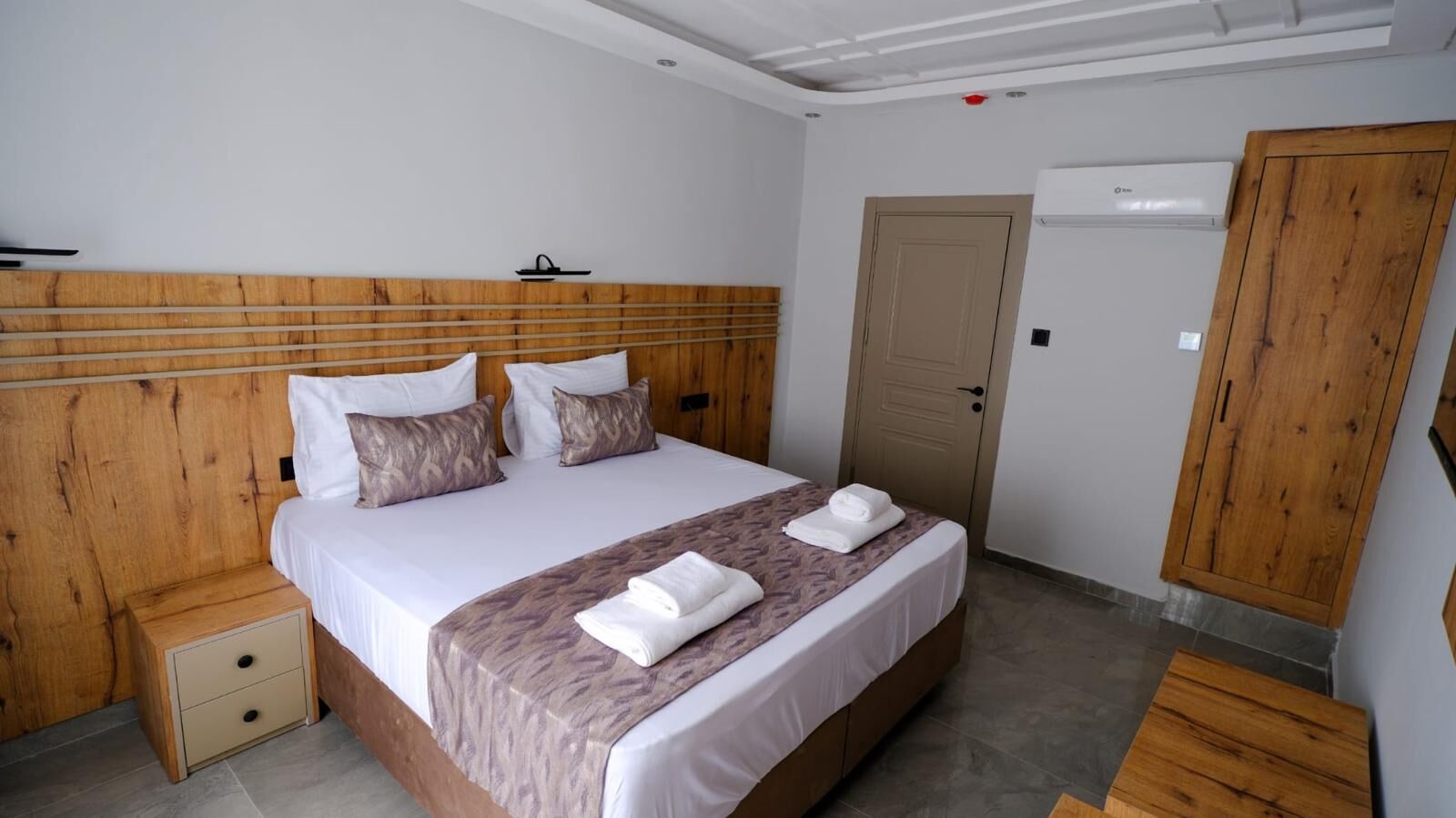 Una habitación de hotel con cabecera de madera, cama con sábanas blancas, mesita de noche de madera y un armario.