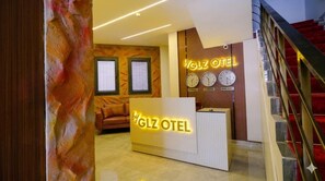 Un mostrador de recepción de hotel con diseño moderno, que incluye una zona de espera, una escalera y relojes mostrando diferentes husos horarios.