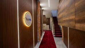 Um corredor de hotel com tapete vermelho, paredes de madeira, uma placa redonda com 'by GLZOTEL' e uma escadaria.