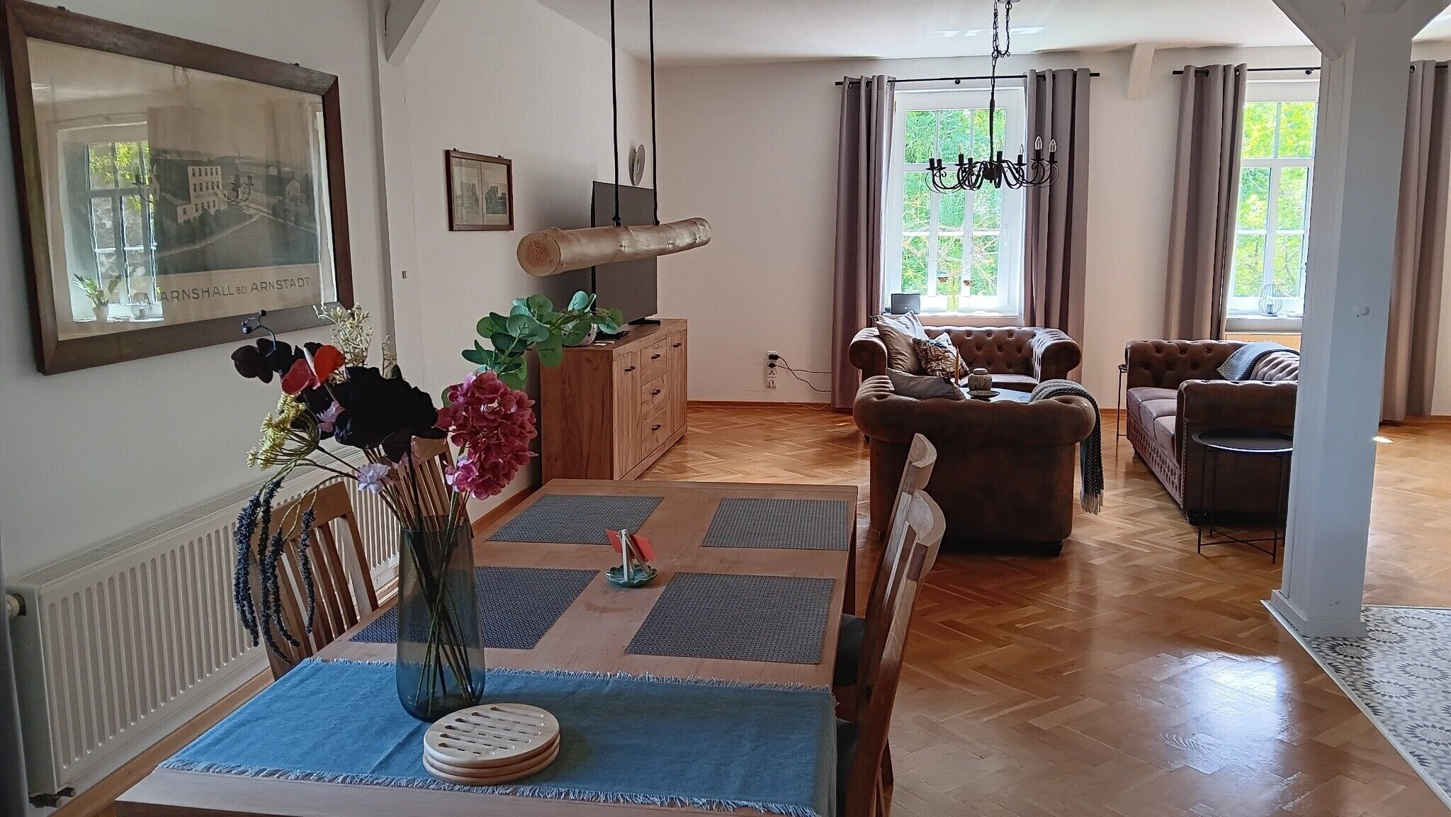 Ein Wohnzimmer mit Holzboden, einem Esstisch mit Blumenvase, einer Couch und einem Sessel.