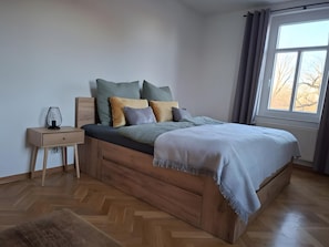 A modern bedroom with a wooden bed frame, a bedside table, and a window with a view of bare trees. - Holiday Apartment Henry – A Break Beneath Historic Beams (Arnstadt)