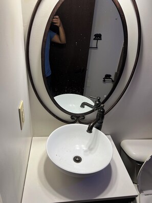 Une salle de bain avec un lavabo blanc, un miroir rond doté d’un cadre noir et une petite décoration représentant un ours noir, accrochée au mur.