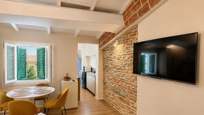 A compact hotel room with a dining area, a TV mounted on the wall, a small kitchen, and a view through a window with shutters.