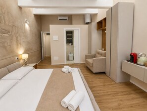 Una habitación de hotel con cama, zona de estar con una silla, una mesita con tetera y un espejo.