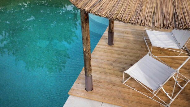Eine Holzterrasse mit Pool, zwei weißen Liegestühlen und einem Reetdachschirm.