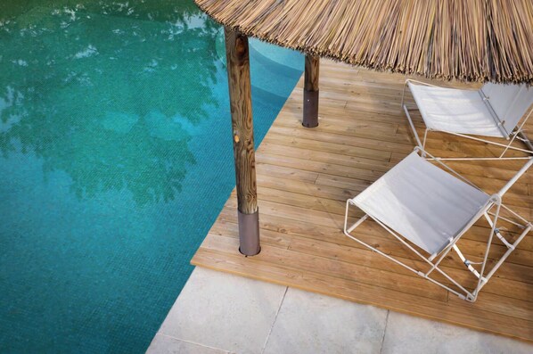 Eine Holzterrasse mit Pool, zwei weißen Liegestühlen und einem Reetdachschirm.