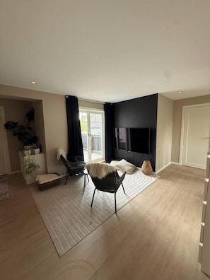 Un salon moderne avec un téléviseur à écran plat, deux fauteuils noirs, une grande fenêtre et une plante.