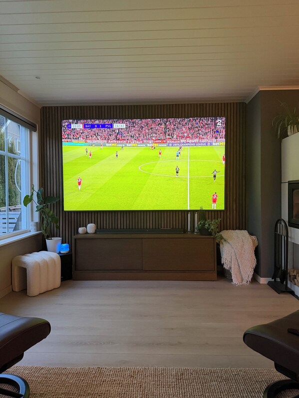 Uma sala de estar com uma televisão grande exibindo uma partida de futebol, um sofá confortável, uma pequena mesa com uma planta e uma janela que proporciona luz natural.