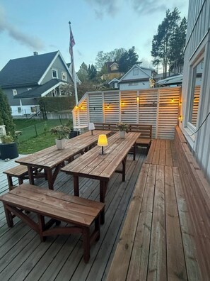 Um deck de madeira com mesas e bancos de piquenique, um candeeiro e uma vedação com luzes de cordão.