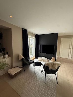 A modern living room with a flat-screen TV, black and white furniture, a large window, and a door.