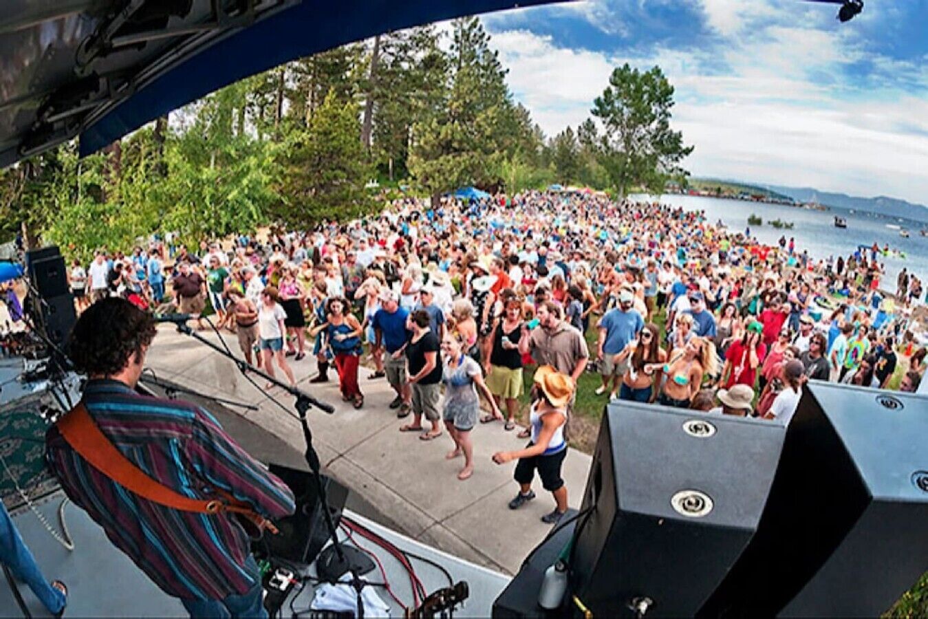 A large outdoor gathering with a stage and equipment, surrounded by trees and a body of water.