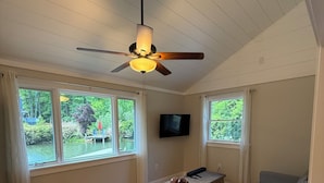 Una habitación con suelo de madera, ventilador de techo, televisor, sofá y ventanas grandes con cortinas.