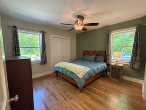 Quarto com cama, ventoinha no teto, cômoda e janela com cortinas.