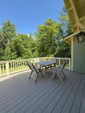 Um deck de madeira com mesa e cadeiras, rodeado por um corrimão branco, com vista para uma área arborizada.