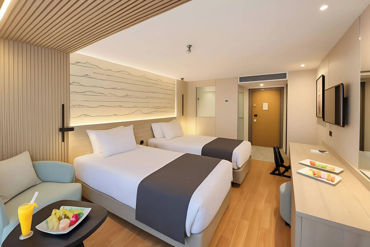 Ein Hotelzimmer mit zwei Betten, einem Schreibtisch, einem Sessel, einem kleinen Tisch mit Obst und einem Fernseher an der Wand.