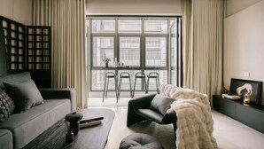 Ein modernes Wohnzimmer mit Sofa, Couchtisch, Sessel, einem großen Fenster mit Vorhängen und einer TV-Konsole mit Bilderrahmen.