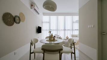 A modern living room with a round dining table, white chairs, a flat-screen TV mounted on the wall, and a large window with sheer curtains.