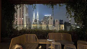A rooftop terrace with a view of the city skyline, including the Petronas Towers, and furnished with wicker seating and metal stools.