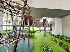 Ein Spielplatz mit einer Seilbrücke, einer runden Kletterkonstruktion und Rasen aus Kunstgrün.