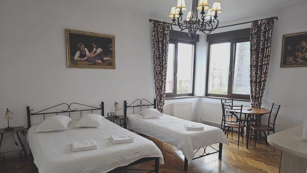Una habitación con dos camas, una mesa de comedor, un candelabro y dos ventanas con cortinas.