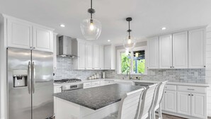 Una cocina moderna con armarios blancos, electrodomésticos de acero inoxidable, encimera de granito e iluminación colgante.