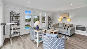 Amplia sala de estar con un sofá gris, sillones estampados, una mesa de centro y un televisor. Hay estantes con objetos decorativos y un ventanal con vistas al exterior.
