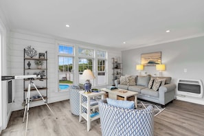 A spacious living room with a grey sofa, patterned armchairs, a coffee table, and a TV. There are shelves with decorative items and a window with a view of the outdoors.