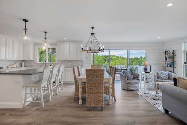 A modern kitchen with white cabinets, a dining area with wicker chairs, and a large window offering a view of greenery.