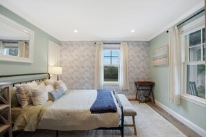 Quarto com cama, mesa de cabeceira, luminária, janela com cortinas e vista para o verde lá fora.