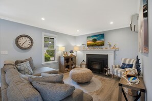 Uma sala de estar com sofá, banquinho, lareira e um quadro na parede.