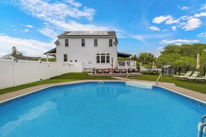 A swimming pool with a white house in the background, a fenced yard, and a clear sky.