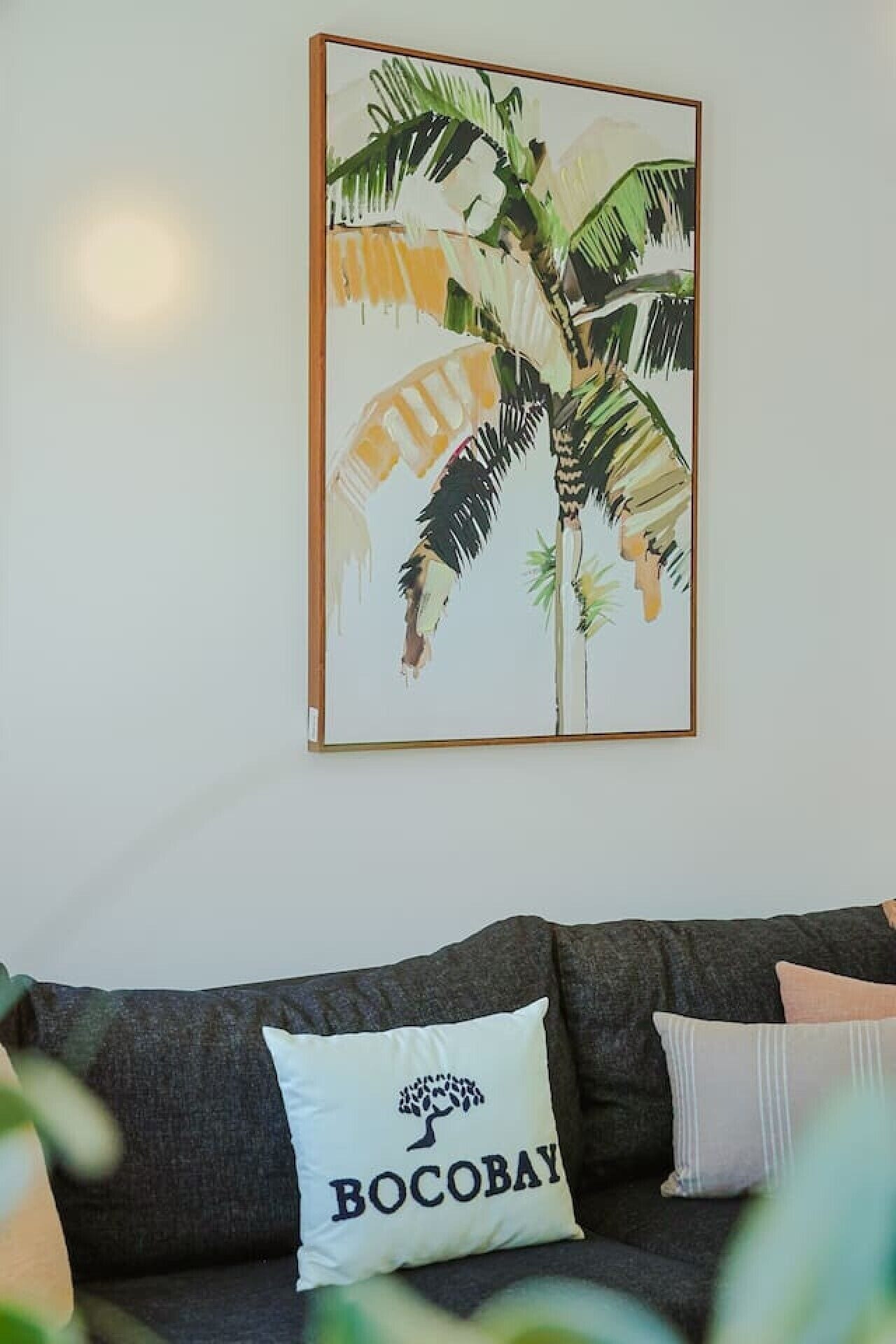 A living room with a framed tropical plant artwork, a dark grey sofa, and a pillow with a tree and text.