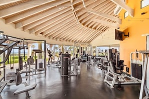 Ein Fitnessstudio mit verschiedenen Trainingsgeräten, einer Holzbalkendecke und Blick auf Palmen.