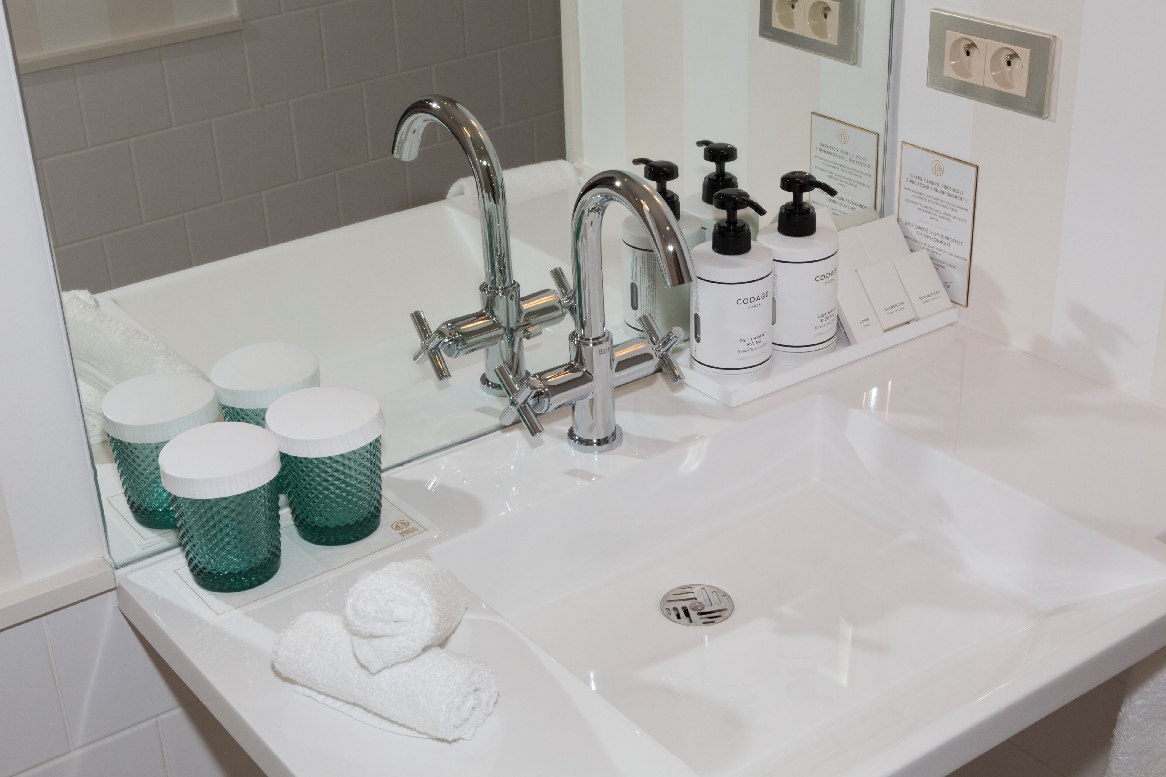 Une salle de bain avec un lavabo blanc, un robinet chromé, des distributeurs de savon verts et un miroir.
