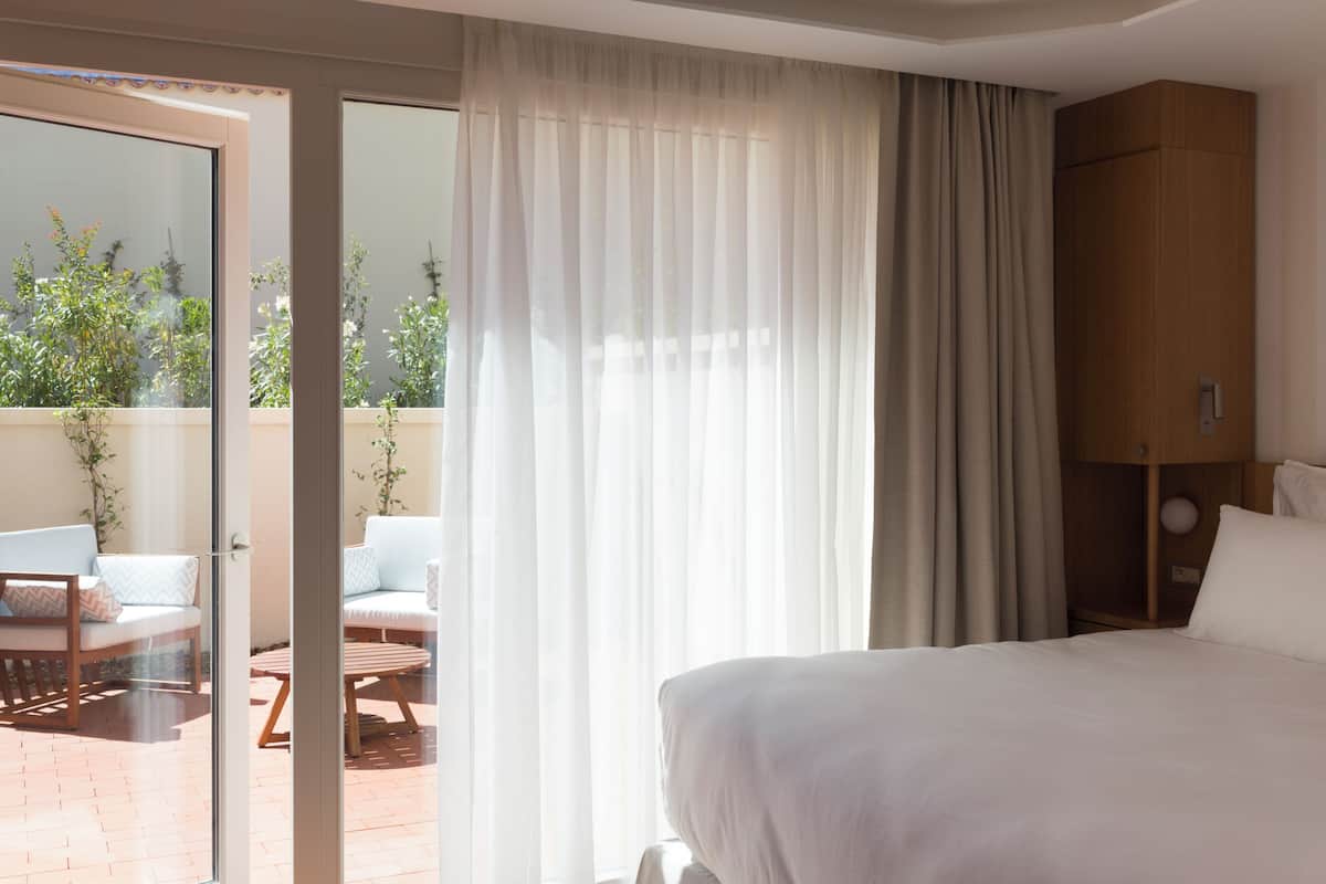 Habitación de hotel con cama, armario de madera, puerta de vidrio que da a un área exterior con mesa y sillas, y vista a la vegetación.