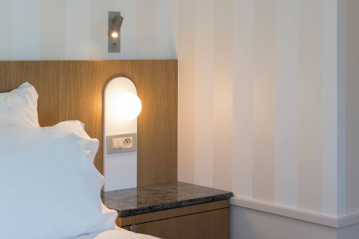Habitación de hotel con cama, mesita de noche, lámpara de pared y cabecero de madera.