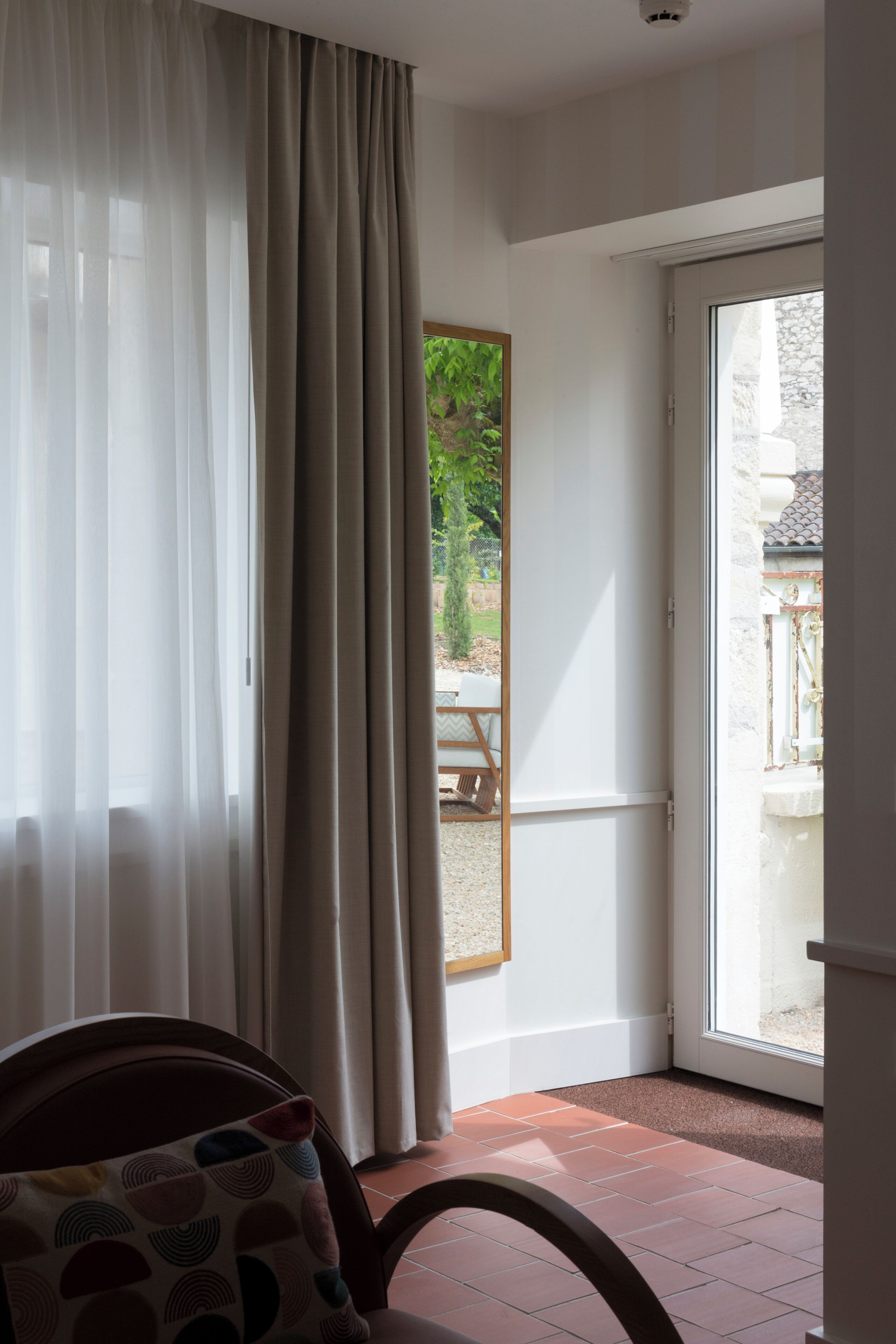 Una habitación con un ventanal grande, una silla con cojín estampado y vistas a un espacio exterior con mesa y sillas.