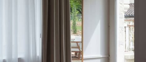 Quarto com uma janela grande, uma cadeira com almofada estampada e vista para área externa com mesa e cadeiras.