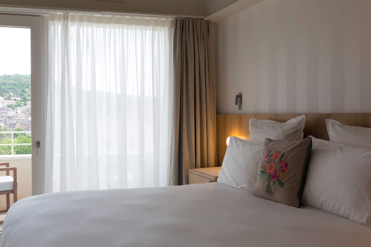 Una habitación de hotel con cama, mesita de noche, lámpara y vistas a la ciudad por la ventana.