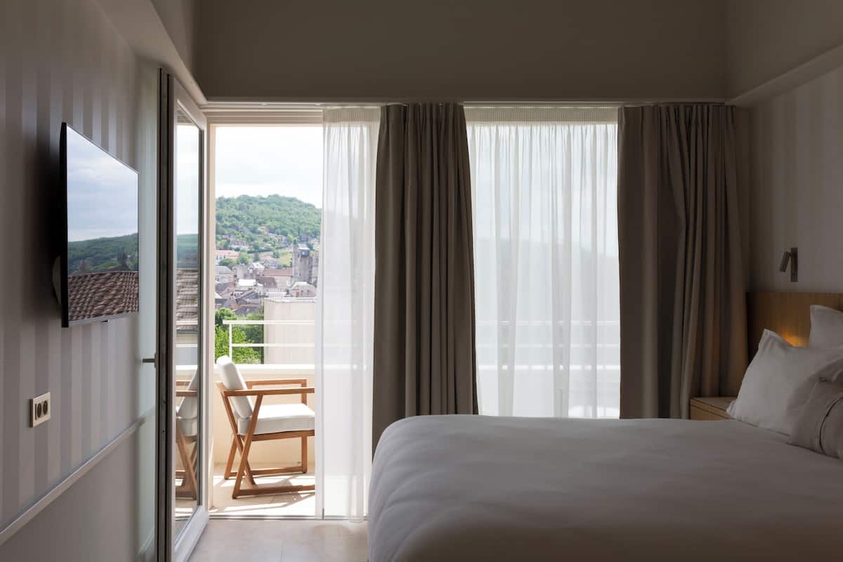 Habitación de hotel con cama, televisor, balcón con una silla y vista a la ciudad.