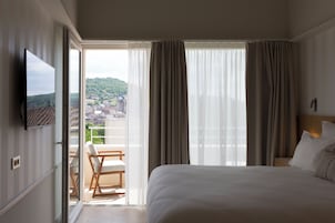 A hotel room with a bed, a TV, a balcony with a chair, and a view of a cityscape.