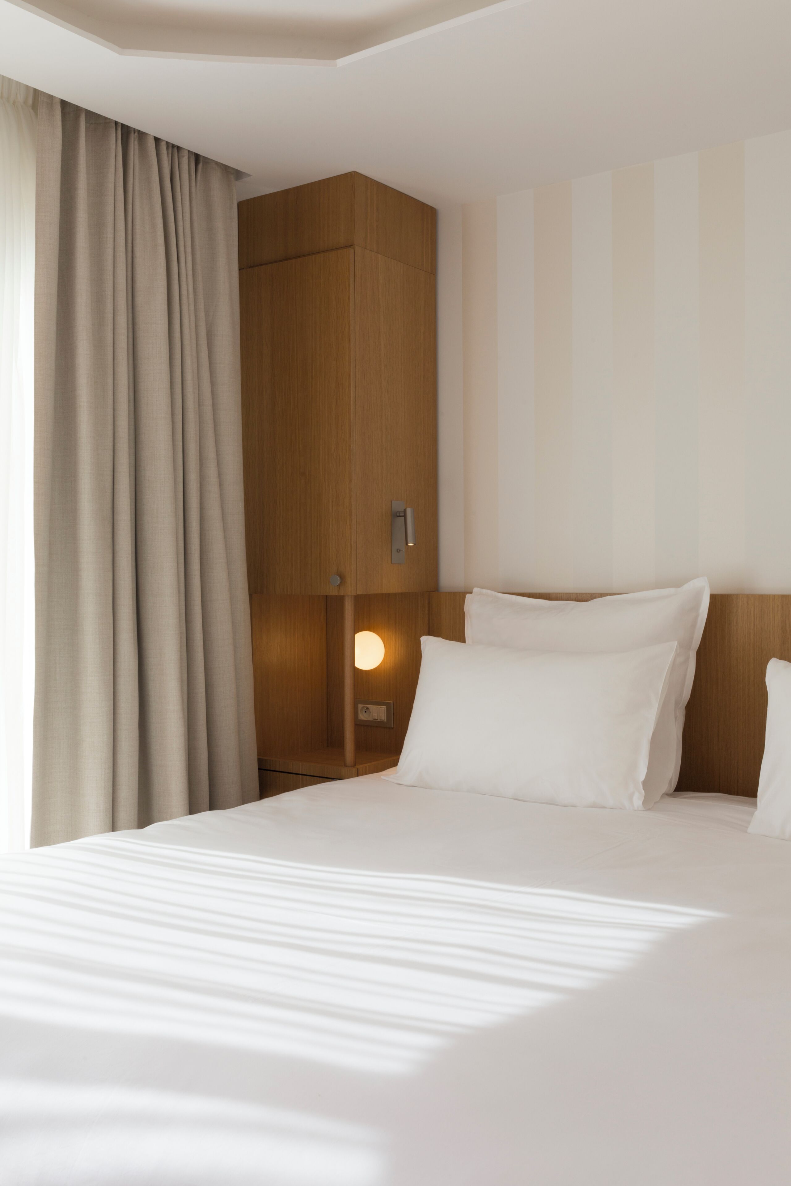 A hotel room with a bed, curtains, a nightstand, and a wall-mounted light.