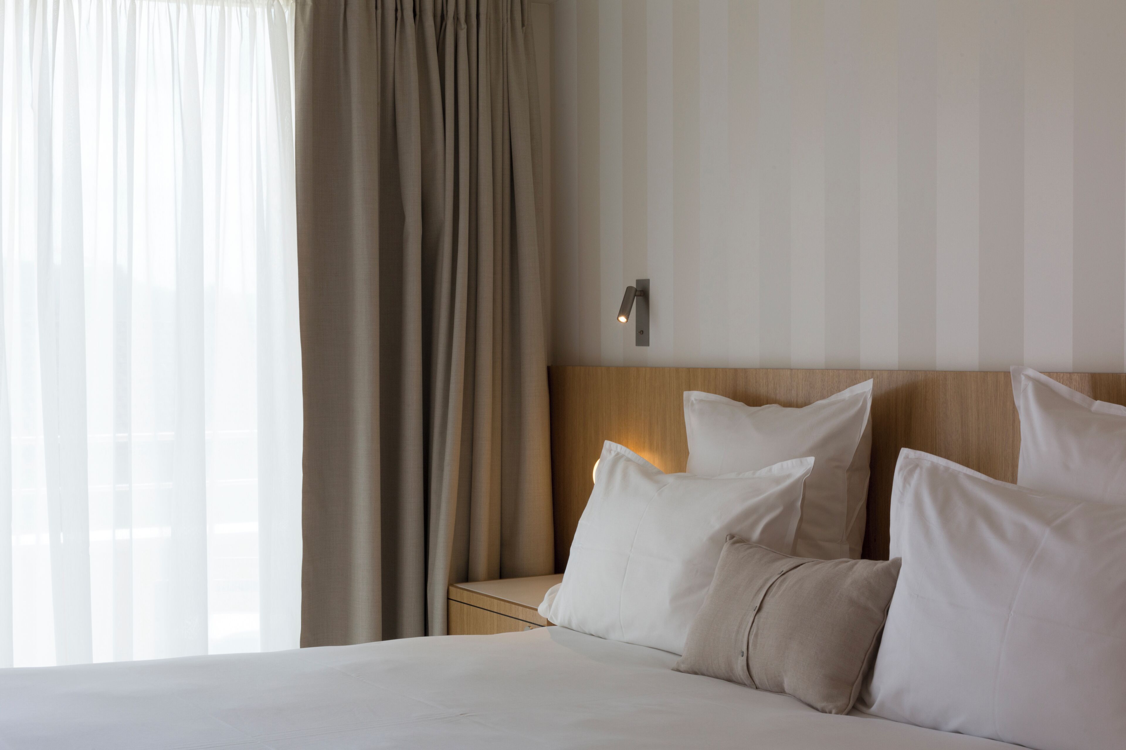 A hotel room with a bed, curtains, a nightstand, and a wall-mounted light.